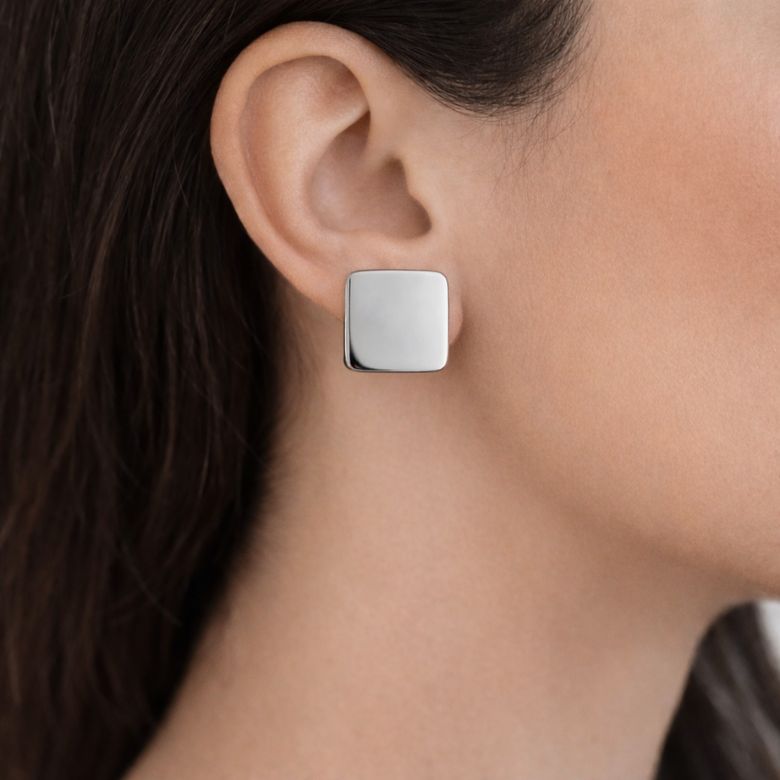 Close-up of silver, square Orecchini Quadrati Lucidi in Acciaio Argentato earring on ear, reflecting light.